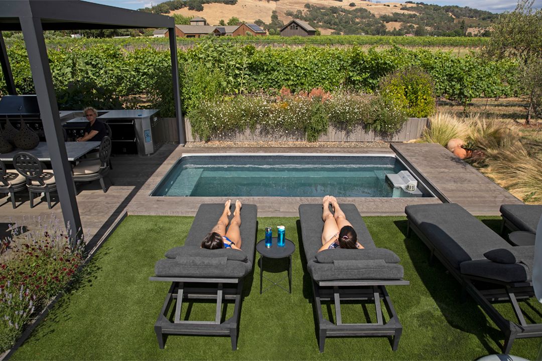 A picture of a pool deck with a partially in-ground swimming pool, tables, chairs, and potted plants.